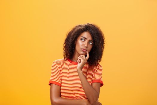 Woman considering to take part in event. Smart focused and creative good-looking african girl with curly hair holding finger on lip gazing concerned left, thinking making decision over orange wall.