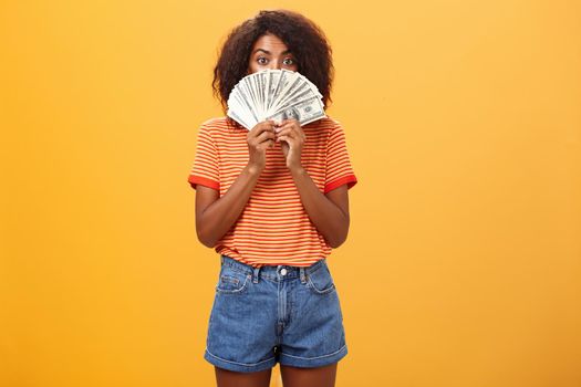 Woman gonna waste lots of cash today. Pleased and delighted rich african american girl with curly hairstyle hiding face behind bunch of money peeking mysteriously at camera winning millions. Copy space