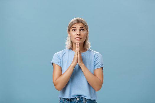 Studio shot of hopeful focused dreamy girl with attractive face and blonde hair holding hands in pray near body looking up hopefully with faith praying or making wish over blue background. Body language concept