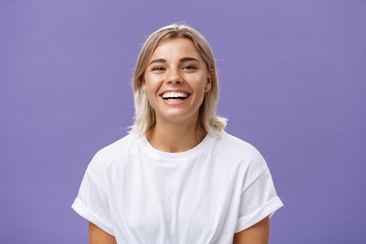 Close-up shot of joyful charming blonde female with delighted and pleased smile standing in white t-shirt over purple background spending time in awesome amusing company. Emotions concept