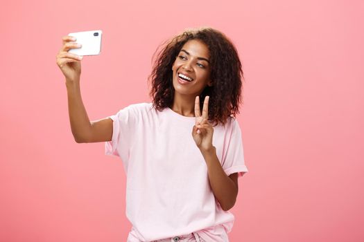 Stylish sociable good-looking dark-skinned female student with curly hairstyle pulling hand with smartphone near face taking selfie showing peace sign to device screen while smiling carefree. Lifestyle.