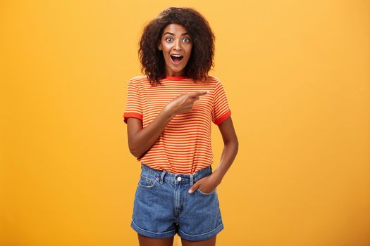 Hey awesme copy space there. Portrait of impressed and surprised enthusiastic young african american female with afro hairstyle pointing right astonished looking energized and curious over orange wall.