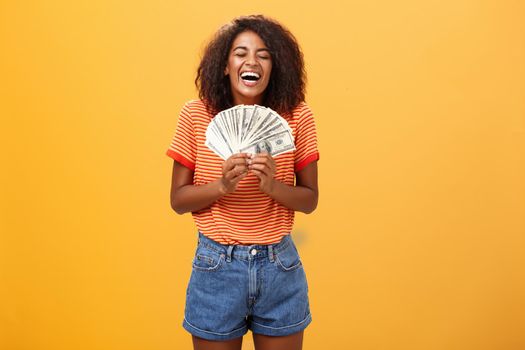 Finally girl rich. Portrait of happy charismatic stylish young female model receiving first payment holding lots of money and laughing from joy winning lottery ready waste cash over orange background.