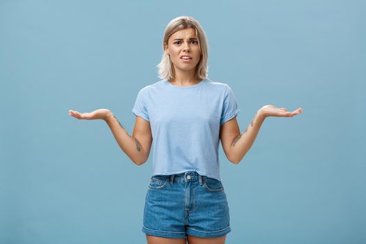 Girl feeling struggles to understand what happening. Confused perplexed intense good-looking female with fair hair shrugging with hands spread aside in displeased clueless pose posing over blue wall.