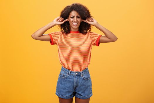 What that anoying sound. Disturbed and displeased dark-skinned girl with curly hair frowning clenching teeth from discomfort closing ears with index fingers from loud irritating sound over orange wall.