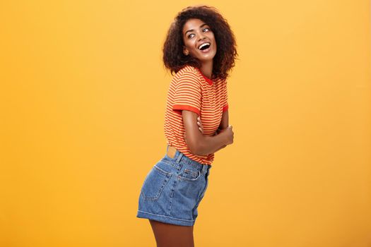 Hey wanna hang out. Sensual and flirty carefree stylish dark-skinned woman with curly hair standing in profile turning left and posing with happy cute expression smiling and seducing over orange wall.