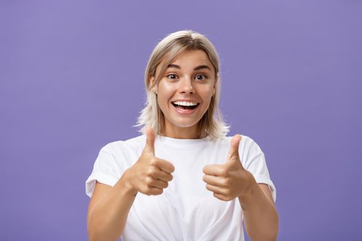 Lifestyle. Waist-up shot of optimistic and supportive good-looking caucasian female in white t-shirt cheering and smiling broadly hearing awesome idea approving it with thumbs up gesture over purple wall.