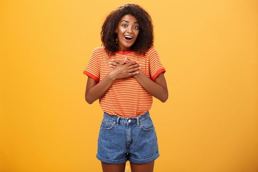 Cute delighted african american woman being grateful and thankful for help standing pleased over orange wall holding palms on breast thanking friend for rescuing grinning joyfully at camera. Lifestyle.