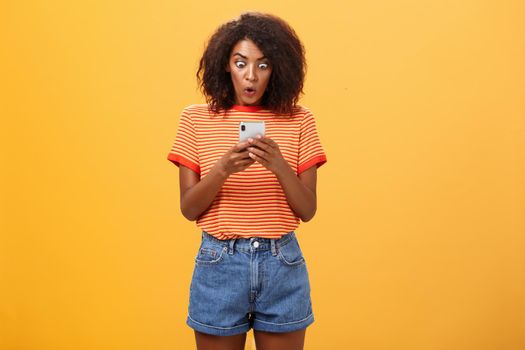 Indoor shot of shocked stunned african american young woman with afro hairstyle staring surprised and excited at smartphone screen holding cellphone reading amazing message over orange wall. Lifestyle.