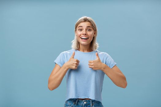 You did great proud of you. Portrait of satisfied and fascinated attractive happy girlfriend in casual t-shirt showing thumbs up and smiling broadly being supportive and cheerful over blue wall.