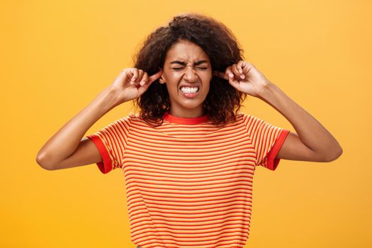 Waist-up shot of intense bothered dark-skinned woman clenchign teeth from discomfort and antipathy closing ears not hear botherind loud noise standing displeased and annoyed over orange wall. Copy space