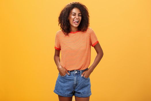 Woman sometimes wants feel childish. Joyful enthusiastic and charismatic young african american female with afro hairstyle winking happily showing tongue holding hands in pockets against orange wall.