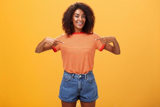 Hey pick me I your girl looking for. Portrait of charming friendly-looking ambitious dark-skinned female with afro hairstyle pointing at chest proudly and joyful posing against orange background.