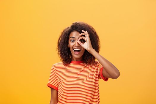 Creative and playful dreamy dark-skinned adult girl with curly hairstyle in striped t-shirt showing circle over eye or okay gesture smiling broadly ready take part in adventures near orange wall. Lifestyle.