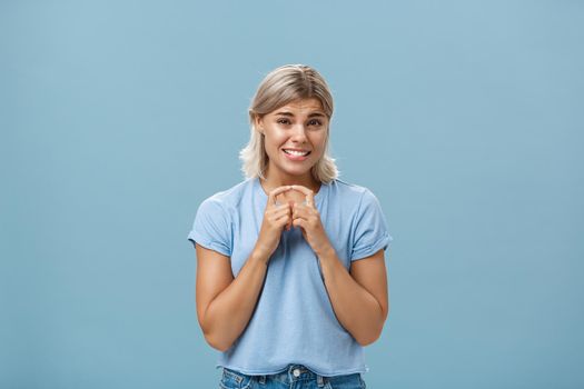Silly cute and worried blonde female friend with short haircut steepling index fingers and smiling with guilty sorry smile frowning while waiting for answer begging for help or favor over blue wall. Copy space