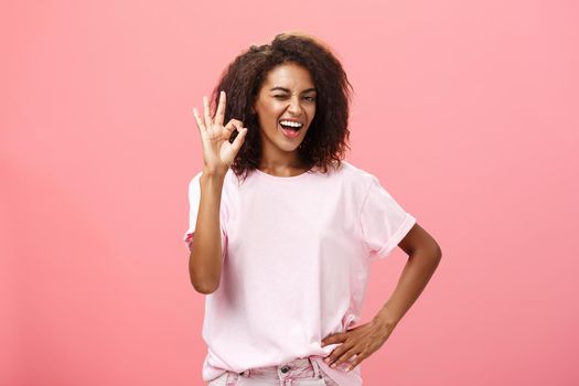 Girl got us covered. Charming charismatic and confident african american skillful woman in stylish outfit holding hand on waist showing okay gesture and winking assured at camera over pink wall.