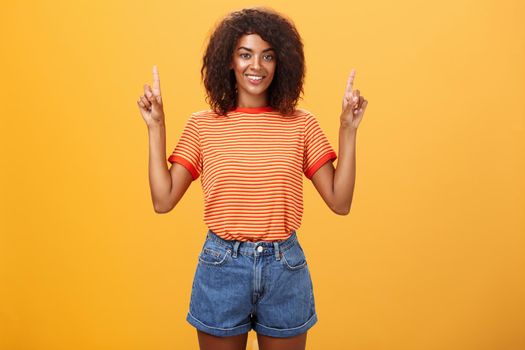 Looking only up and forward. Optimistic ambitious stylish dark-skinned female student in striped cool t-shirt and shorts raising hands pointing upwards and smiling friendly over orange wall.