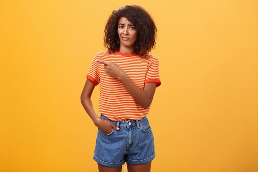 Not worth my time. Displeased unimpressed stylish attractive dark-skinned female model in striped t-shirt and denim shorts frowning with picky expression pointing left doubtful and dissatisfied. Copy space
