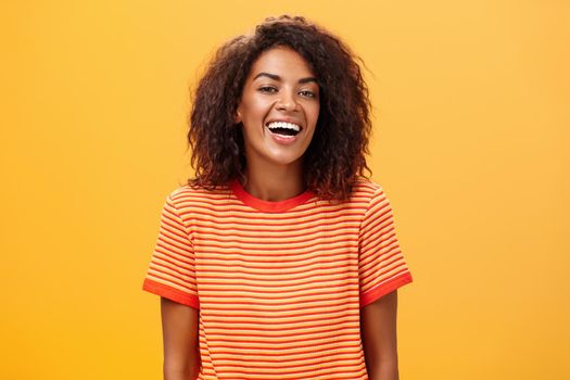 Waist-up shot of outgoing happy charming dark-skinned female with curly hairstyle laughing joyfully posing in striped trendy t-shirt over orange background enjoying nice casual conversation. Lifestyle.