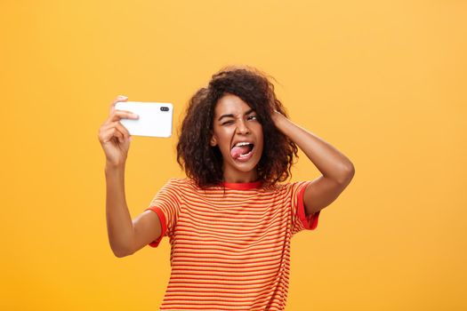 Waist-up shot of stylish confident african american woman with afro hairstyle in trendy t-shirt taking selfie on awesome new cellphone sticking out tongue playing with hair and winking at smartphone. Technology and people concept