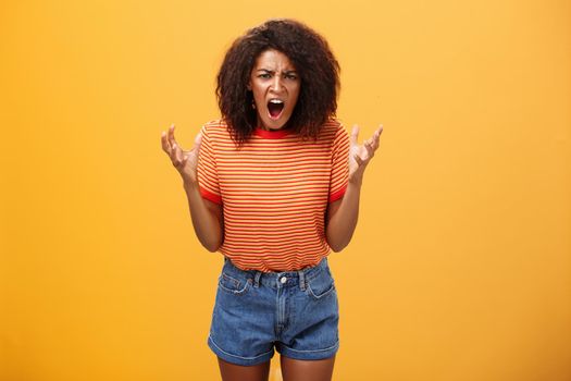 Pissed and outraged angry african american young female in stylish outfit raising palms gesturing and yelling losing temper arguing with cheating partner feeling fed up over orange background. Lifestyle.