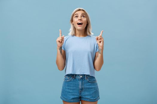 Lifestyle. Indoor shot of impressed speechless attractive fair-haired female student in casual t-shirt and denim shorts dropping jaw from amazement pointing and looking up intrigued over blue wall.