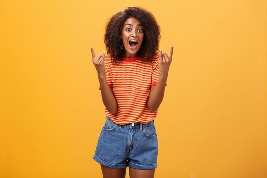 Stylish african american woman being delighted and excited before cool concert showing rock n roll gesture with both hands screaming from satisfaction and happiness cheering over orange background. Copy space