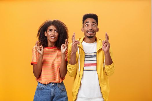 Couple of friends praying and looking up in sky intense crossing fingers for good luck clenching teeth faithfully hoping parents not saw them smoking standing together over orange background. Lifestyle.