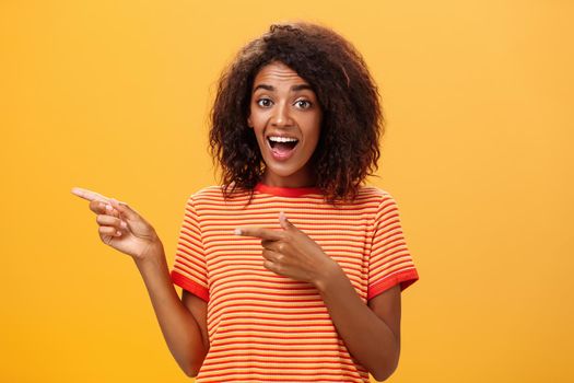 Portrait of amazed excited charismatic dark-skinned young pretty girl with afro hairstyle in trendy striped t-shirt pointing left delighted and fascinated posing against orange background. Lifestyle.