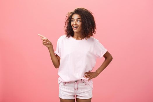 Girl checking out nice body of guy passing by. Charming confident dark-skinned female with curly hairstyle holding hand on hip looking and pointing left discussing cool copy space over pink wall.