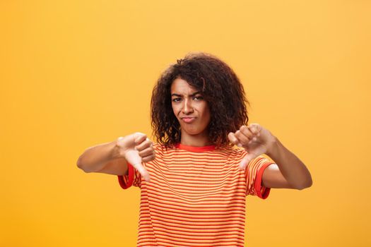 You loser and idea sucks. Dissatisfied and displeased young creative female artist with curly hairstyle frowning, sulking showing thumbd down in dislike gesture giving negative answer over orange wall.