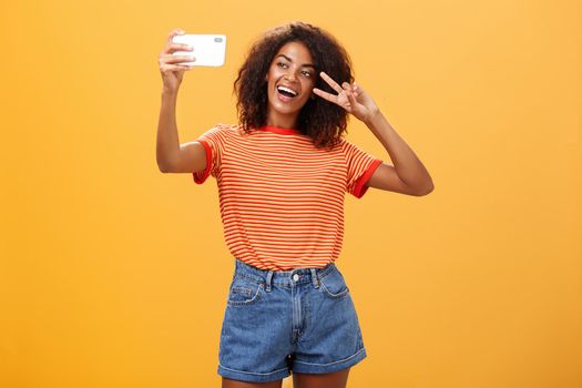 Girl making video vlog with brand new smartphone posting in internet trying become famous standing over orange background posing for selfie gazing at gadget screen showing peace or victory gesture. Copy space