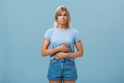 Portrait of displeased intense attractive young blonde girl with tattooes frowning touching stomach complaining on stomachache after eating junk food having disorder over blue background. Health concept