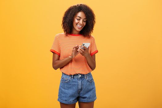 Stylish carefree girl texting friend come over standing pleased over orange wall in stylish denim shorts typing message or scrolling news in internet via smartphone gazing at device screen with smile. Lifestyle.