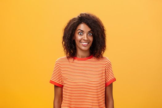 Waist-up shot of amazed and excited charming african american woman with curly hairstyle popping eyes from thrill and joy smiling broadly being surprised by great gift over orange background. Lifestyle.