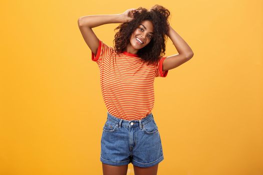 Time start living life fullest. Joyful optimistic woman having fun during vacation tilting head touching curly hair and enjoying summer sunshine in trendy striped t-shirt and shorts over orange wall.