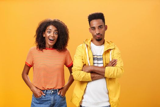 Two african american friends standing over orange background, girl thinks idea is awesome feeling excited and joyful while boyfriend being displeased and indifferent to unimpressive plan. Copy space