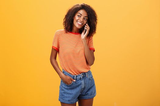 Stylish dark-skinned girl making casual phone call to friend telling all details of after romantic date standing pleased and carefree over orange background in striped t-shirt gazing left with grin. Lifestyle.