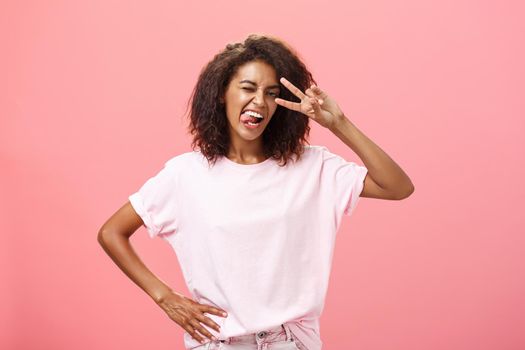 Not afraid express myself. Joyful charismatic african american woman in t-shirt with afro haircut showing tongue playfully and daring making peace sign over eye and winking posing over pink background.