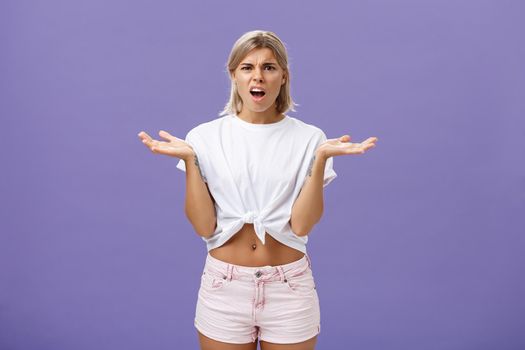 Lifestyle. Perplexed and disappointment good-looking blonde female student in white t-shirt and pink shorts frowning shrugging with spread hands near shoulders saying what the hell over purple wall.