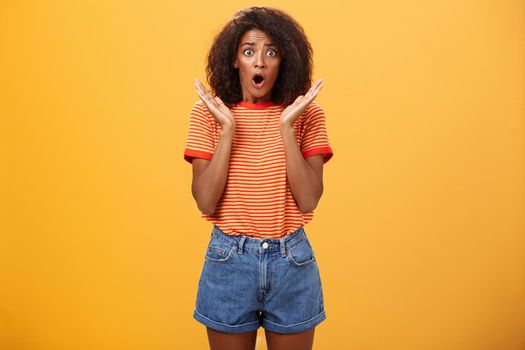 Oh no what happened. Worried african american sister with curly hair in trendy striped t-shirt and denim shorts gasping dropping jaw, looking concerned expressing empathy and shock over orange wall.