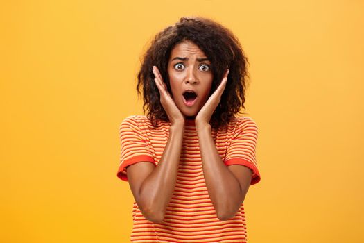 Waist-up shot of shocked concerned panicking african-american woman with afro hairstyle in trendy t-shirt gasping holding palms on face from surprise posing troubled against orange wall. Lifestyle.