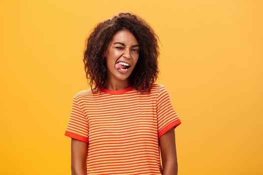 Portrait of daring and emotive confident flirty woman with afro hairstyle winking joyfully showing tongue posing carefree and enthusiastic against orange background flirting with hot guy. Copy space