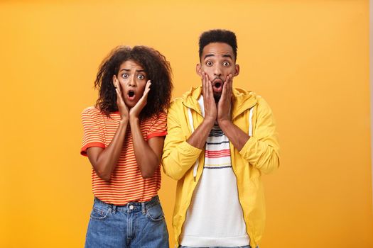 Portrait of shocked and stunned speechless girlfriend dropping jaw from amazement with boyfriend feeling amazed from shook news posing together surprised and astonished over orange wall. Lifestyle.