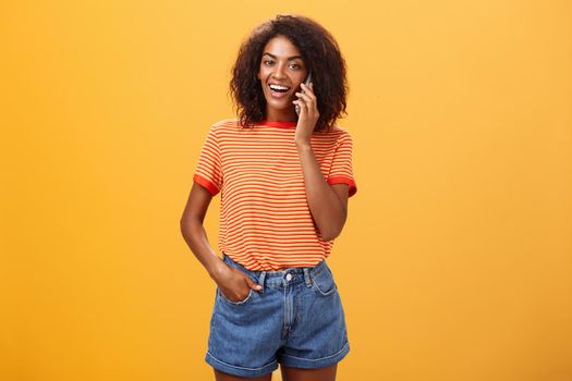 Indoor shot of stylish confident female coworker in trendy t-shirt and denim shorts holding smartphone near ear calling friend talking via cellphone casually about girls stuff over orange background. Lifestyle.