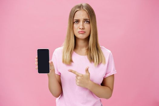 Gloomy and sad cute young female showing friend negative feedback about her project in internet pointing at smartphone screen standing concerned and upset against pink background. Technology and emotions concept