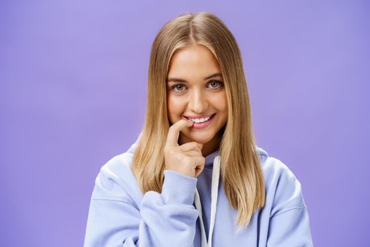 Close-up shot of feminine and tender young girl with fair hair and tanned skin biting finger flirty and shy smiling cute at camera standing in trendy over-sized hoodie, flirting over purple background. Emotions concept