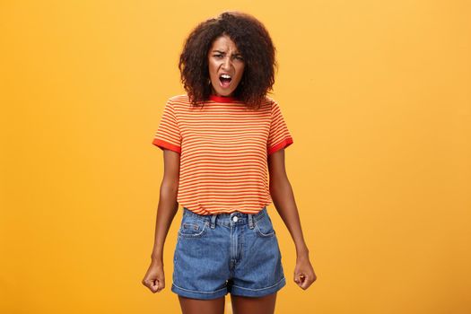 Girl complaining arguing with parents cause acting unfair. Displeased pissed and offended silly african american with afro hairstyle clenching teeth standing in insulted pose yelling over orange wall.