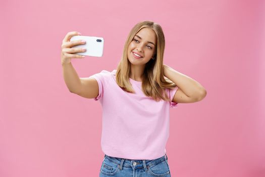 Female beauty blogger taking selfie post new look online. Portrait of charming tanned young woman in t-shirt touching hair gently pulling hand with smartphone near face taking photo over pink wall. Technology concept