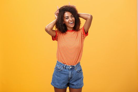 Rebellious playful and joyful african american girl with curly hairstyle making devil horns on head with index fingers smiling broadly showing daredevil character and charisma over orange wall. Lifestyle.